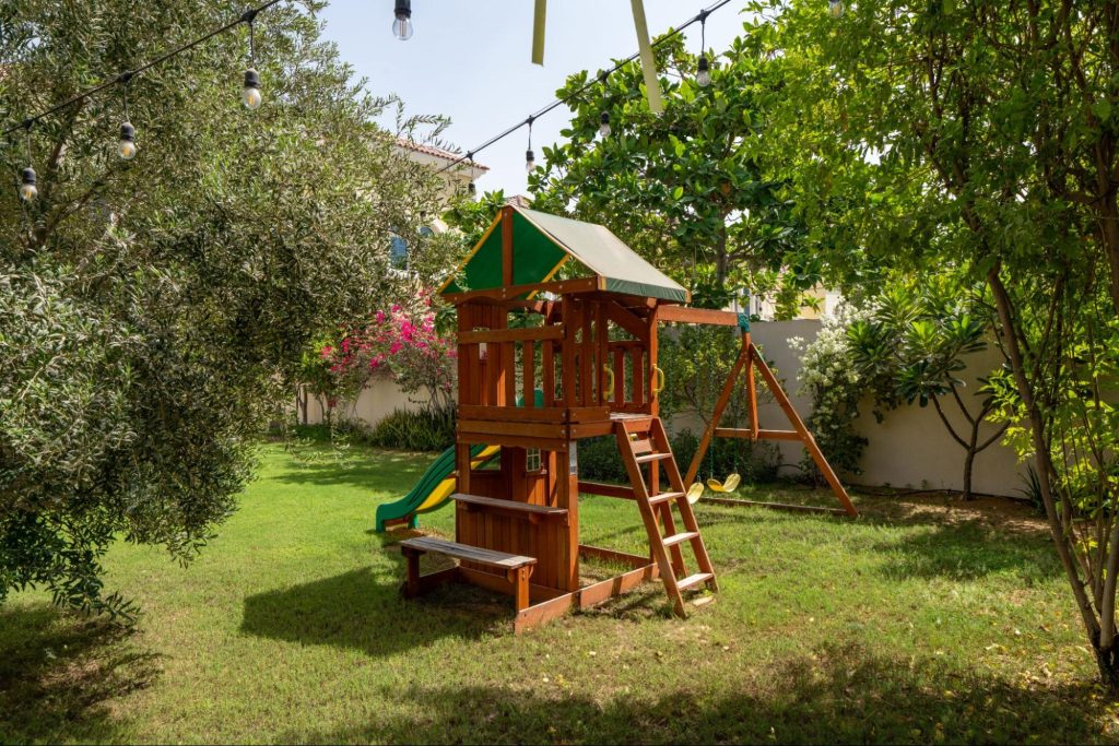 Children’s garden design by Smart Renovation for Casa Fiorita featuring a play area with a wooden swing set, surrounded by lush trees and soft lawn.